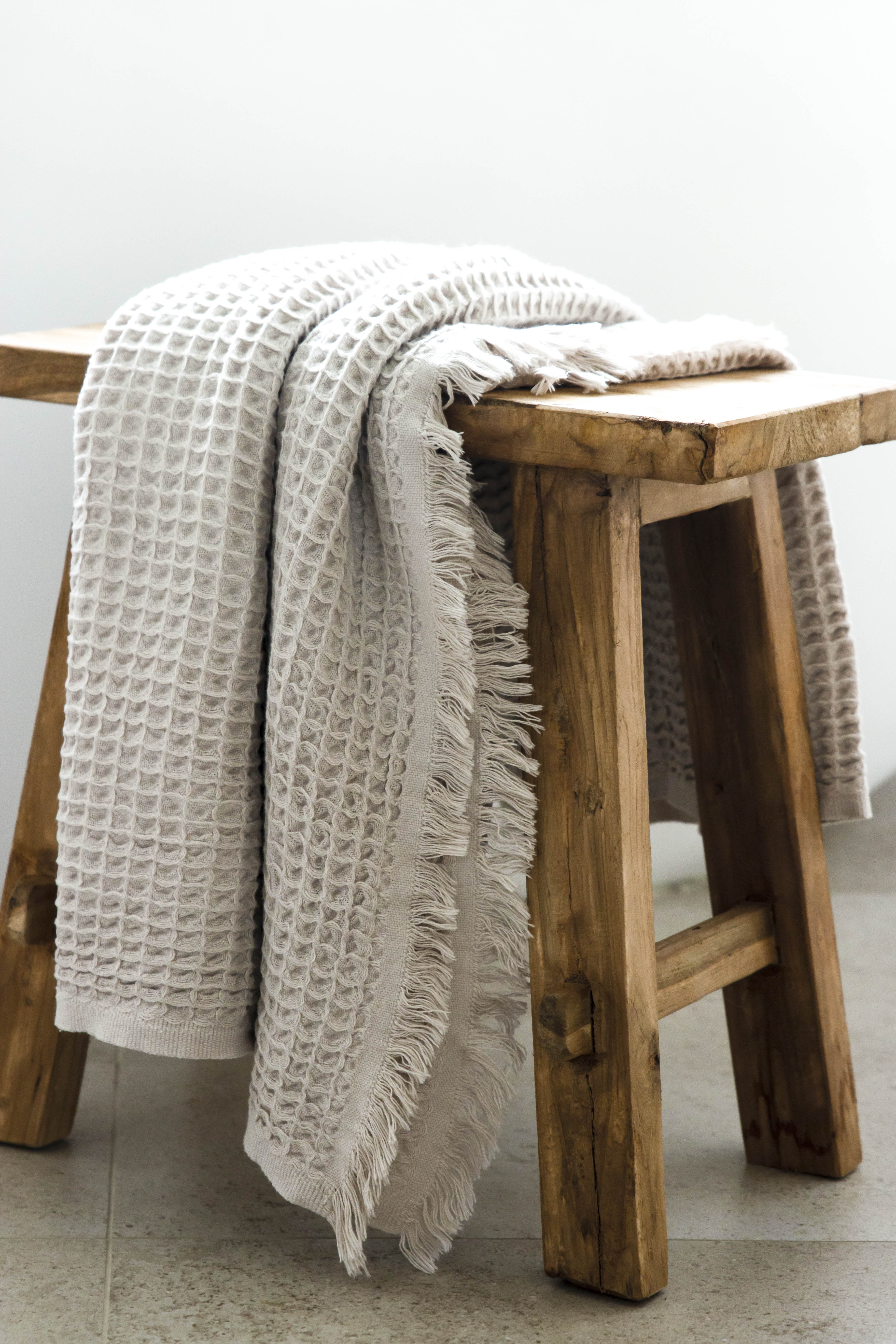 An oatmeal coloured waffle bath towel with tassles, lying over a wooden stool.