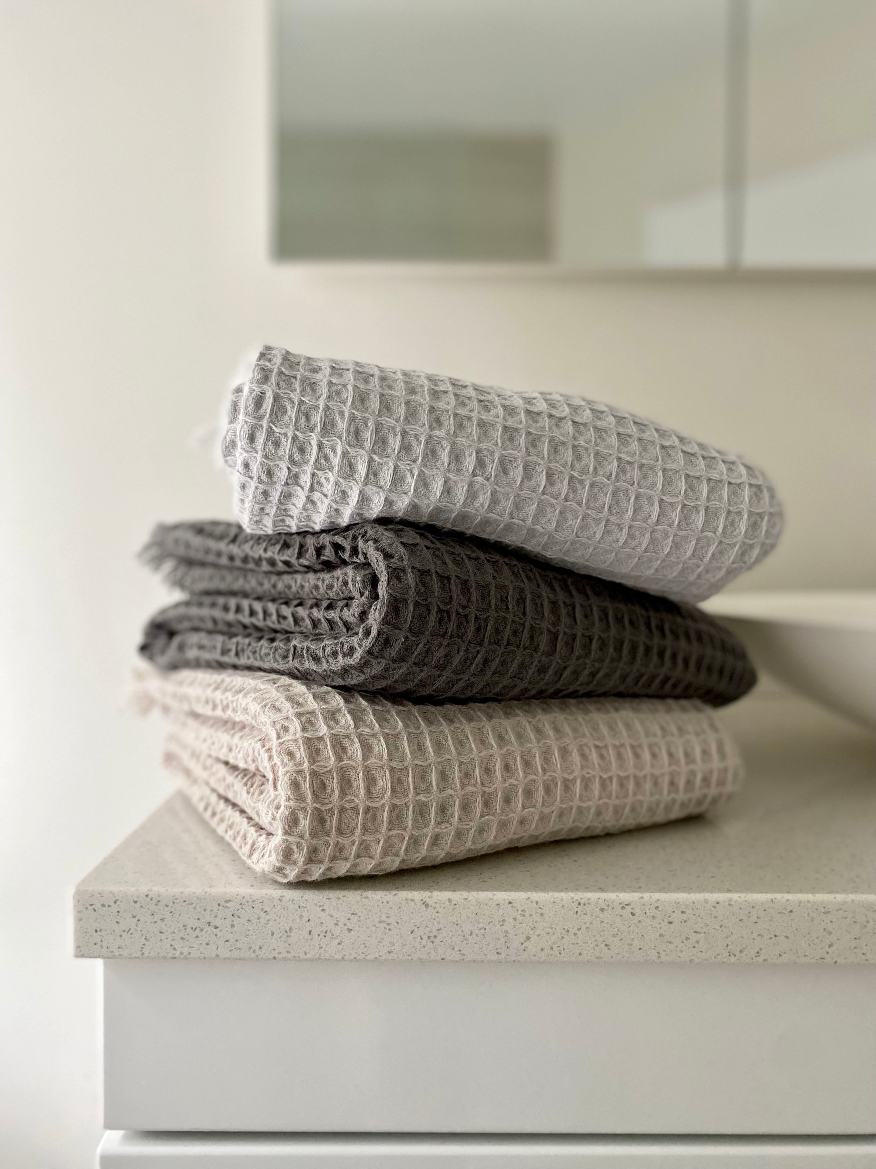 3 folded waffle bath towels in pale grey, stonewash and oatmeal on a bathroom bench