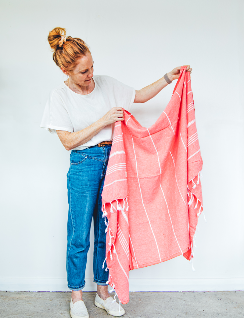 a lady holds up a watermelon Turkish towel with white stripes, displayed vertically with the fringes at the bottom for display