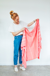 a lady holds up a watermelon Turkish towel with white stripes, displayed vertically with the fringes at the bottom for display